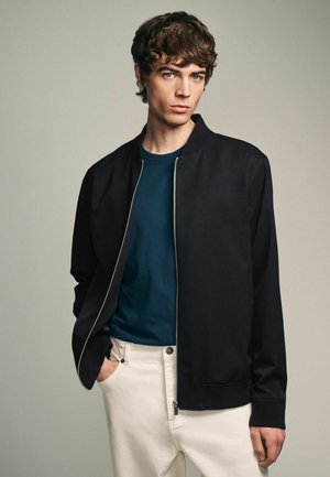 Chaqueta bomber negra con cierre frontal, puños acanalados y bolsillos, llevada sobre una camiseta de color verde oscuro, combinada con pantalones blancos. Textura suave.