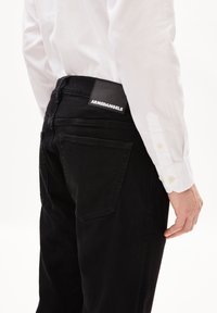 Des jeans en denim noir avec des poches arrière et une étiquette carrée avec le logo "ARMEDANGELS". Associés à une chemise blanche à manches longues. Texture lisse.