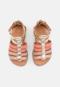 Kaksi nyöritettyä sandaalia, joissa on yhdistelmä vaaleanpunaista ja metallista nauhaa, tekstuuripintainen muotoilu ja sileä ruskea pohja. Solki sivussa.