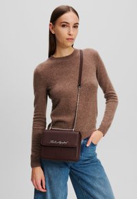 Maglione marrone a maglia, jeans blu e una piccola borsa a tracolla in bordeaux con texture, dotata di una cinghia in metallo argentato e dettagli con logo.