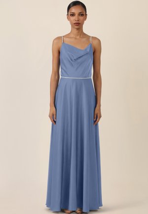 Vrouw staand, gekleed in een lichtblauwe, mouwloze jurk tot op de grond met dunne bandjes en een gedrapeerde halslijn, tegen een beige achtergrond.