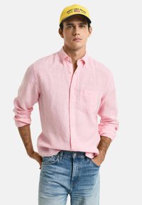 Lichtroze linnen overhemd met knoopjes en een borstzak, opgerolde mouwen, gecombineerd met vervaagde blauwe jeans. Model draagt een gele pet.
