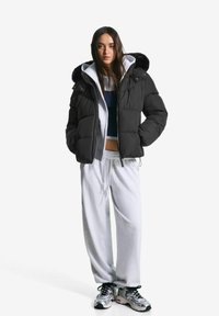 Vrouw staat in een zwarte pufferjack met een bontcapuchon, grijze hoodie, losse witte sweatpants en zilveren sneakers op een witte achtergrond.