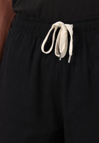 Pantalons noirs à coupe relaxée en tissu texturé, avec une taille élastique et un cordon crème avec des embouts en métal argenté.