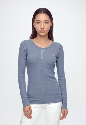 Donna con capelli lisci e scuri indossa una camicia blu aderente a maniche lunghe con bottoni e pantaloni bianchi con dettagli ricamati.