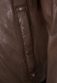 Braune Lederjacke mit glatter Textur, dezentem Glanz und einem knopflosen Akzent auf der Tasche. Das Design ist tailliert mit klaren Linien.