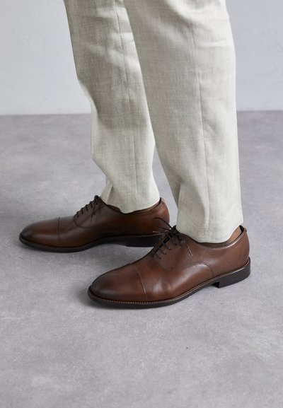 Chaussures de ville en cuir marron avec un design épuré, bout rond et fermeture à lacets, accompagnées de pantalons ajustés de couleur claire.