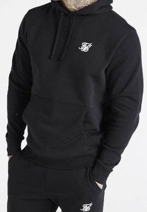 Person trägt schwarzen Hoodie und schwarze Jogginghose, beide mit weiß gesticktem Logo, Hände in den Taschen, sichtbarer tätowierter Hals.