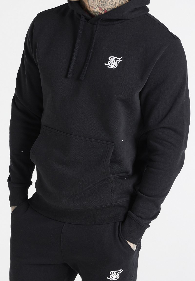 Personne portant un hoodie noir et un pantalon de survêtement noir, tous deux avec un logo brodé blanc, mains dans les poches, cou tatoué visible.