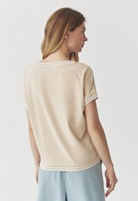 Beige lyhythihainen paita, jossa on rento leikkaus, pyöreä kaula-aukko ja raidallinen reunus hihoissa ja helmassa, vaaleaa taustaa vasten.
