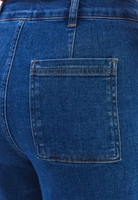 Tmavě modré džíny s zadní kapsou, zdobené oranžovým prošíváním a hladkou strukturou. Materiál vypadá odolně a má klasický design.