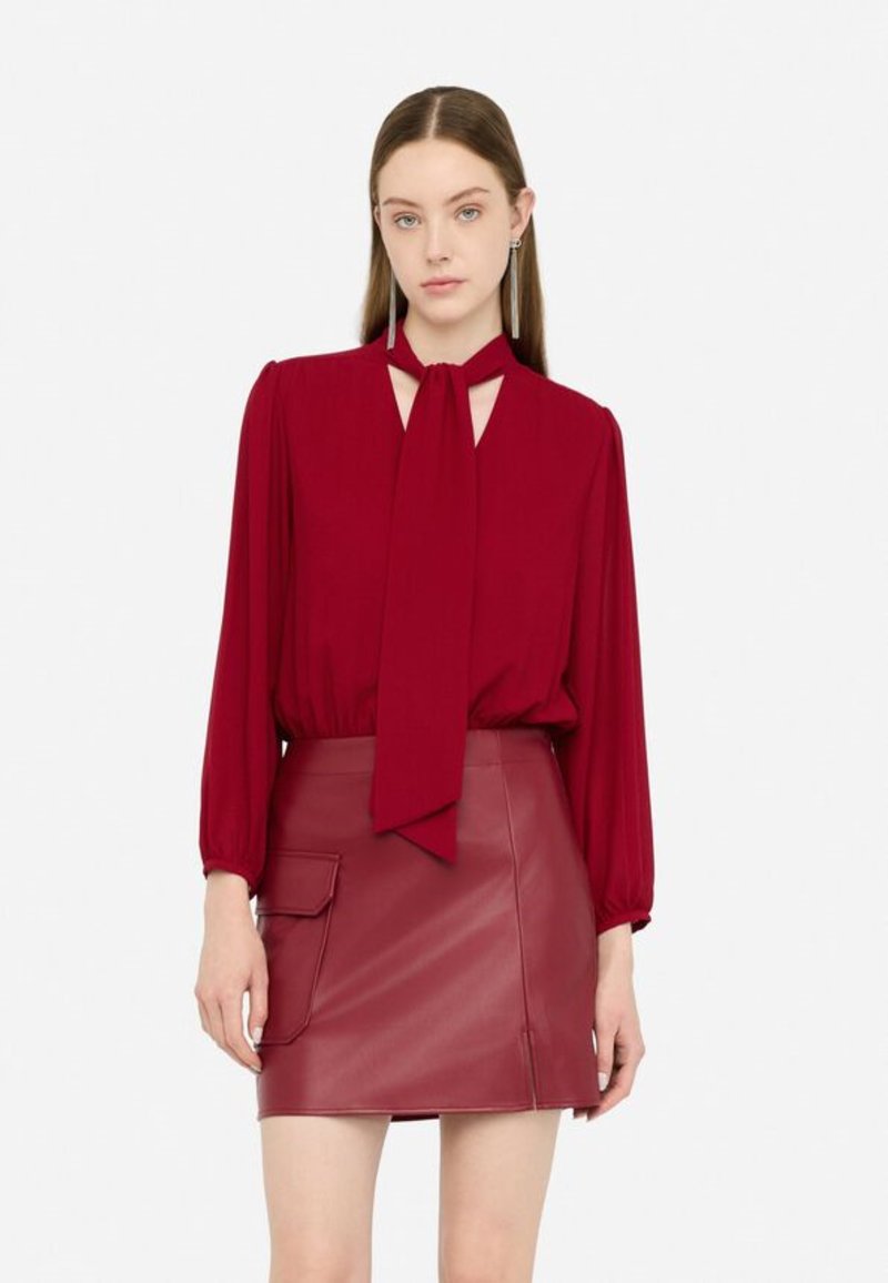 Blusa marrone con maniche lunghe e colletto a lacci, abbinata a una mini gonna in pelle coordinata con tasca laterale. Texture morbida.