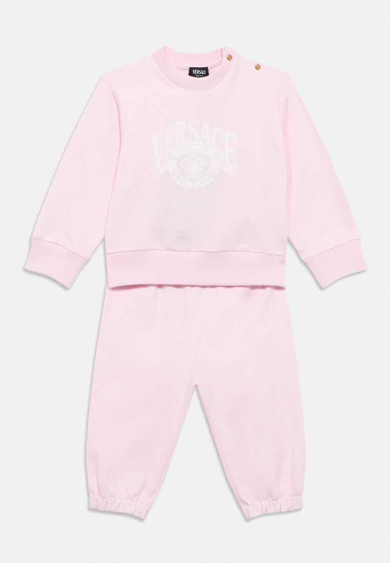 Σετ ρούχων από απαλό ροζ ύφασμα, που περιλαμβάνει φούτερ με στρογγυλή λαιμόκοψη και λευκό λογότυπο Versace, καθώς και τζόκερ παντελόνια με ελαστικές μανσέτες.