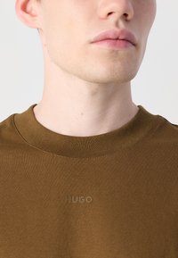 Braunes Baumwoll-T-Shirt mit strukturierter Oberfläche, ausgestattet mit einem gerippten Rundhalsausschnitt und einem geprägten "HUGO"-Logo auf der Brust. Nahaufnahme.