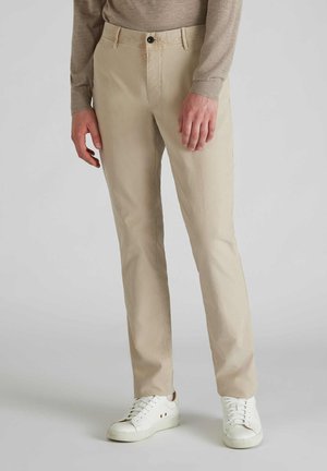 Beige Baumwoll-Chinos mit geradem Schnitt, ausgestattet mit einem Frontknopfverschluss und Seitentaschen, kombiniert mit weißen Sneakers.