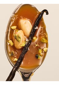Cuillère avec sauce caramel, amandes entières, pistaches concassées, flocons de sel marin et gousse de vanille sur fond beige.