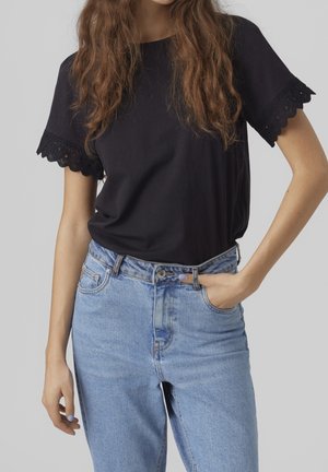 Femme portant un haut noir à manches courtes avec bordure en dentelle et un jean bleu clair taille haute, une main dans la poche.