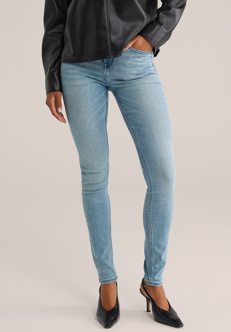 Žena oblečená v svetlo modrých skinny džínsach, čiernej koženej bunde a čiernych vysokých podpätkoch s otvorenou zadnou stranou.