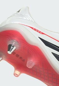 Λευκή σόλα παπουτσιού ποδοσφαίρου με κόκκινες λεπτομέρειες, με υφή επιφάνειας, μαύρες ρίγες και ασημένιες μεταλλικές τάπες για πρόσφυση.