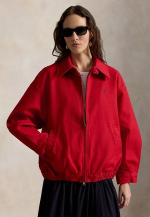 Kobieta w czarnych okularach przeciwsłonecznych i czerwonej kurtce na zamek, z rękami w kieszeniach na neutralnym tle.