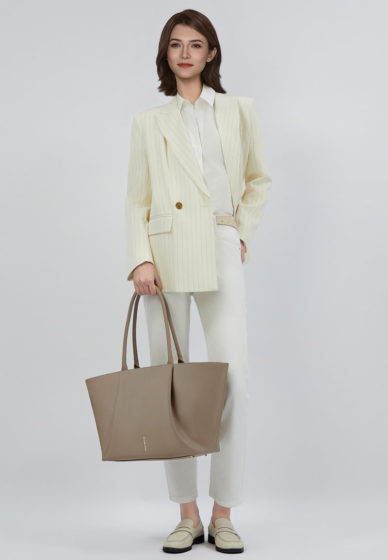 Vrouw in een crèmekleurige pinstripe blazer, witte blouse en broek, beige loafers, die een grote taupe schoudertas vasthoudt tegen een effen achtergrond.