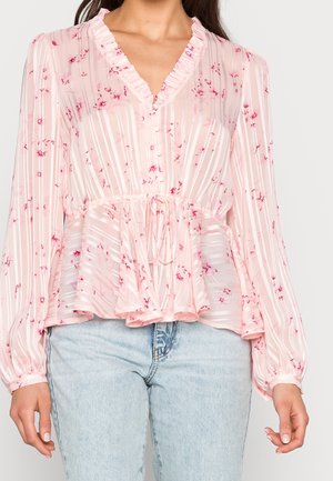 Kvinde iført en lyserød semi-gennemsigtig bluse med blomsterprint, flæsekant, knapper, snøre i taljen og lyseblå jeans.