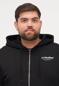 Zwarte zip-up hoodie van zacht materiaal, met een voorlogo "JJ Studios" in het wit. Het heeft een trekkoordcapuchon en een standaard pasvorm.