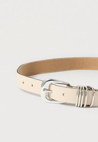 Δερμάτινη ζώνη σε κρεμ απόχρωση με λείο φινίρισμα. Διαθέτει ασημένια μεταλλική αγκράφα και τρεις διακοσμητικούς μεταλλικούς κρίκους στο λουράκι.