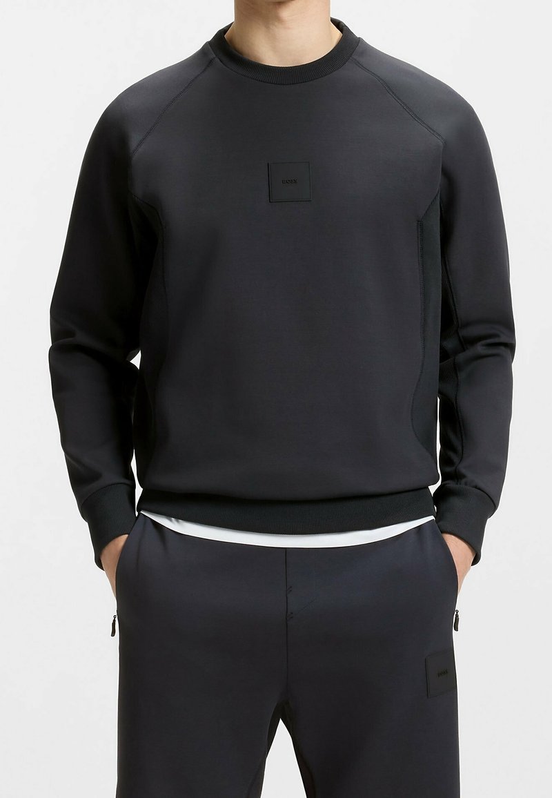 Homme portant un sweat-shirt noir à manches longues et un pantalon noir assorti, les mains dans les poches, debout devant un fond uni.
