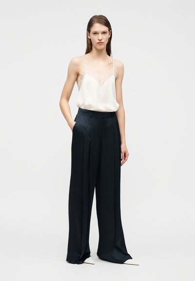 Νεαρή γυναίκα που στέκεται με τα χέρια στις τσέπες, φοράει λευκή μπλούζα με δαντελωτή λεπτομέρεια και φαρδιά μαύρη παντελόνα με λευκά παπούτσια.