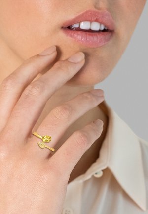 Goldring mit einem gelben Edelstein und einem blattförmigen Akzent. Glatter Ring und polierte Oberfläche, getragen an einer Hand in der Nähe des Gesichts.