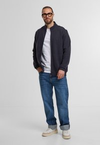 Marineblauer Zip-Up-Bomberjacke mit hohem Kragen, getragen über einem weißen T-Shirt, kombiniert mit lässigen blauen Jeans und beigen Sneakers.