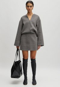 Grauer Wollmischung-Oberteil mit V-Ausschnitt und weiten Ärmeln, kombiniert mit einem passenden Rock. Das Model hält eine Tasche aus schwarzem Lackleder und trägt schwarze kniehohe Stiefel.
