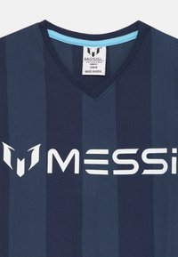 Námornícky modrý dres s V-čkovým výstrihom a vertikálnymi tmavými pruhmi; obsahuje bielu logu "MESSI" a emblém; vyrobený z mäkkej tkaniny. Nálepka: "VYROBENÉ V INDII."