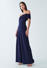 Tamsiai mėlynas romperis be petnešėlių su sukryžiuota krūtinės dalimi, platėjančiomis kelnėmis ir glotnia tekstūra. Auksiniai aukštakulniai su dirželiais papildo aprangą.