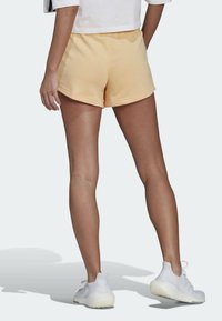 Ljust gula bomullsshorts med en rundad nederkant, parat med vita träningsskor. Slät textur och avslappnad passform, fotograferat bakifrån.