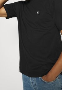 Camiseta negra de manga corta hecha de algodón, con un pequeño diseño bordado en el lado izquierdo del pecho y un corte holgado. Combinada con jeans de mezclilla azul.