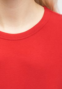 Κοντινό πλάνο λαιμού και άνω μέρους στήθους που φοράει απλή κόκκινη μπλούζα με στρογγυλή λαιμόκοψη, με εμφανές ανοιχτόχρωμο δέρμα και ανοιχτόχρωμα μαλλιά.