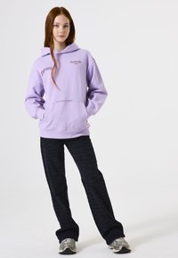 Adolescente con cabello rojo largo que lleva una sudadera ligera morada con texto, pantalones negros con estampado y zapatillas grises, de pie con las manos en los bolsillos.