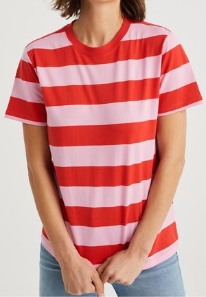 Vrouw die een rood-roze T-shirt met horizontale strepen en korte mouwen draagt, en lichtblauwe jeans, staand tegen een effen achtergrond.