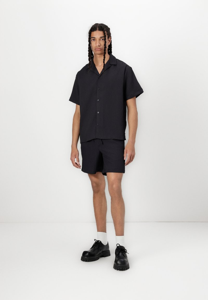 Kortærmet sort tekstureret skjorte med knapper, parret med matchende shorts, suppleret med sorte sko og hvide sokker. Minimalistisk design.