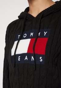 Crna pletena dukserica s kapuljačom s uzorkom pletenice, koja ima pravokutni logo u crvenoj, bijeloj i plavoj boji s natpisom "TOMMY JEANS" u bijelim slovima.