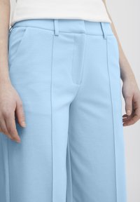 Gai zilu plati bikses, kas izgatavotas no mīksta auduma, ar priekšējiem krokām, sānu kabatām un piegrieztu jostasvietu strukturētai pieguvei.