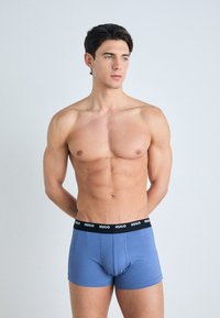 Blaue Baumwoll-Boxershorts mit einem schwarzen Bund, der das "HUGO"-Logo trägt. Das Modell steht mit den Armen hinter dem Rücken und hat einen neutralen Gesichtsausdruck.