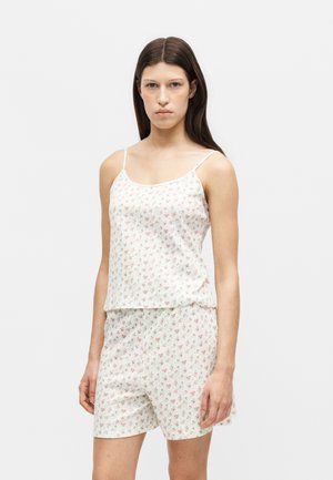 Femme aux longs cheveux foncés portant un haut blanc à fines bretelles fleuri et un short assorti, debout devant un fond clair uni.