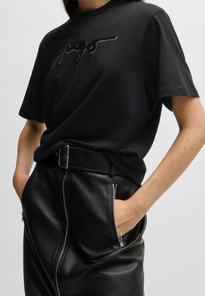 Person iført en sort nittet t-shirt med logo, stukket ned i et sort lædernederdel med lynlås foran og lynlåslommer, hænderne i lommerne.