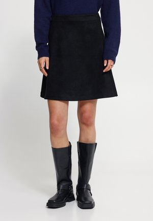 Femme portant une jupe noire au genou, un pull bleu foncé et des bottes en cuir noires montant jusqu'aux genoux, debout sur un fond blanc.