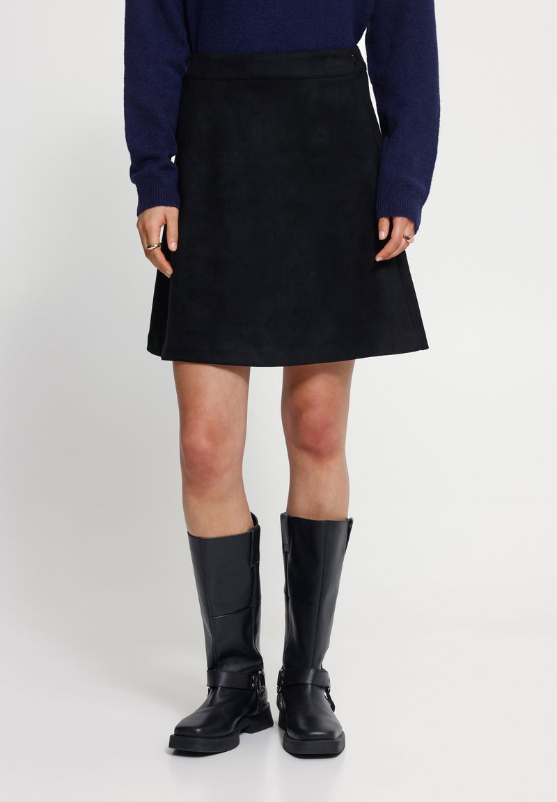 Femme portant une jupe noire au genou, un pull bleu foncé et des bottes en cuir noires montant jusqu'aux genoux, debout sur un fond blanc.