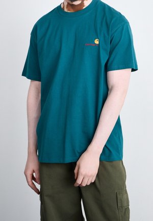 Mężczyzna w turkusowej koszulce Carhartt i oliwkowych spodenkach typu cargo, z rękami luźno opuszczonymi wzdłuż ciała na jednolitym tle.