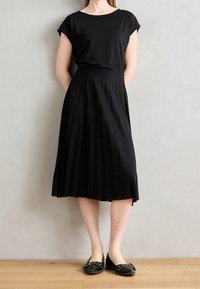 Musta lyhythihainen paita yhdistettynä laskostettuun mustaan midi-hameeseen, joka esittelee sileää pintaa. Mallilla on mustat ballerinat puulattialla.